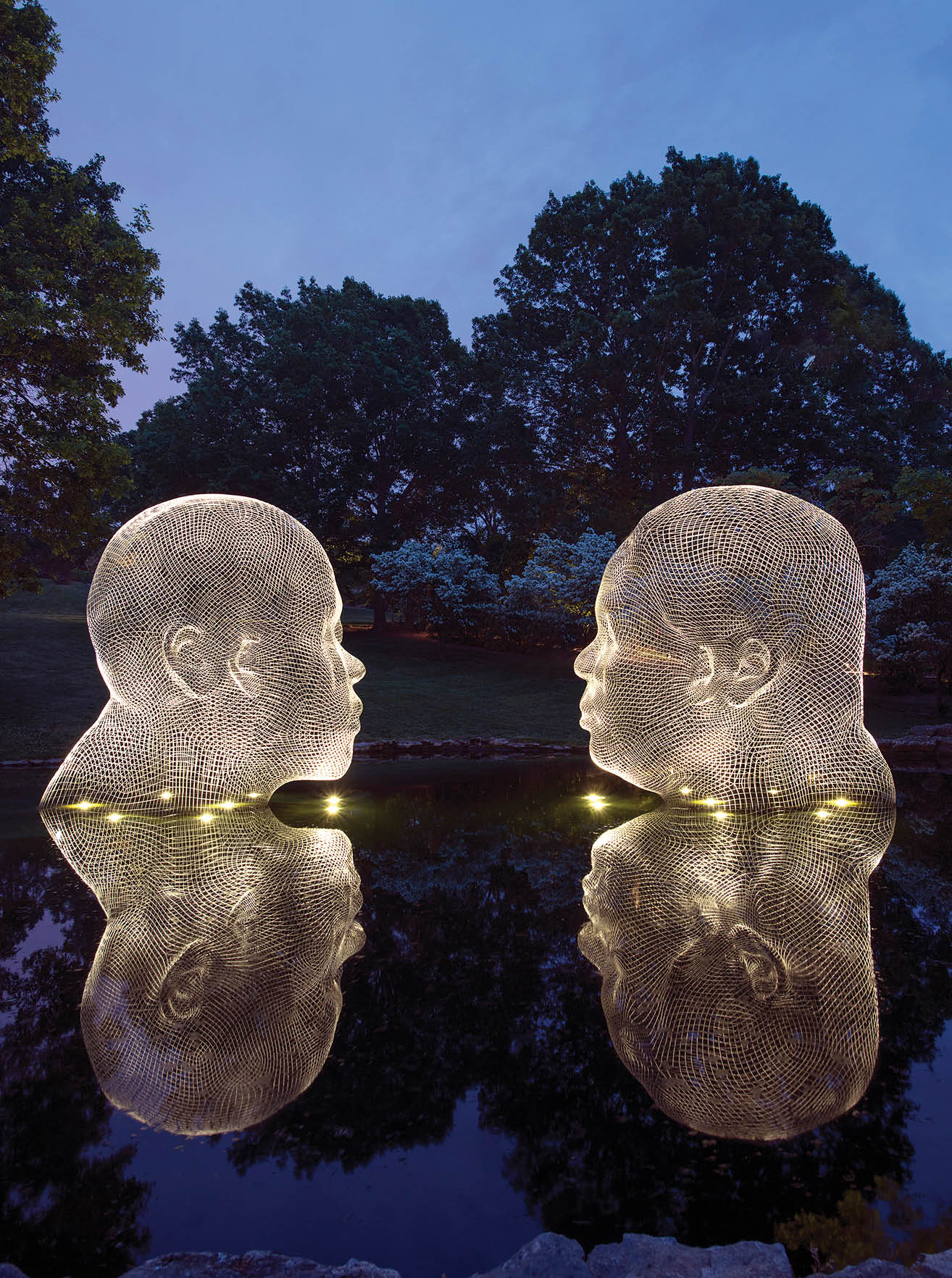 Two illuminated sculptures of human heads are displayed in a pond, facing each other. AI generated content