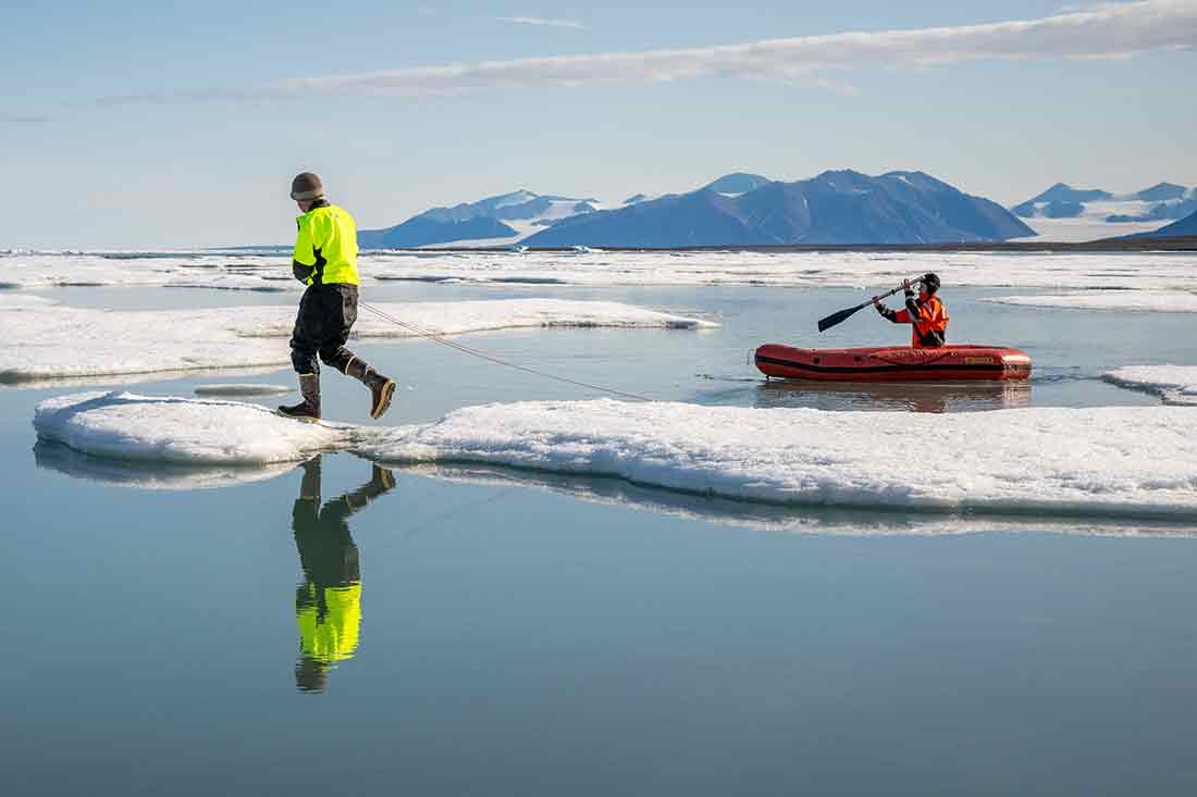 Brian Buma and Alejandra Borunda work to cross a small lead over open water between the mainland and the Arctic Sea icepack, beginning the trek to the northernmost land in the world, Inuit Qeqertaat (Kaffeklubben Island). One person walking on snow and another paddling in an inflatable boat.