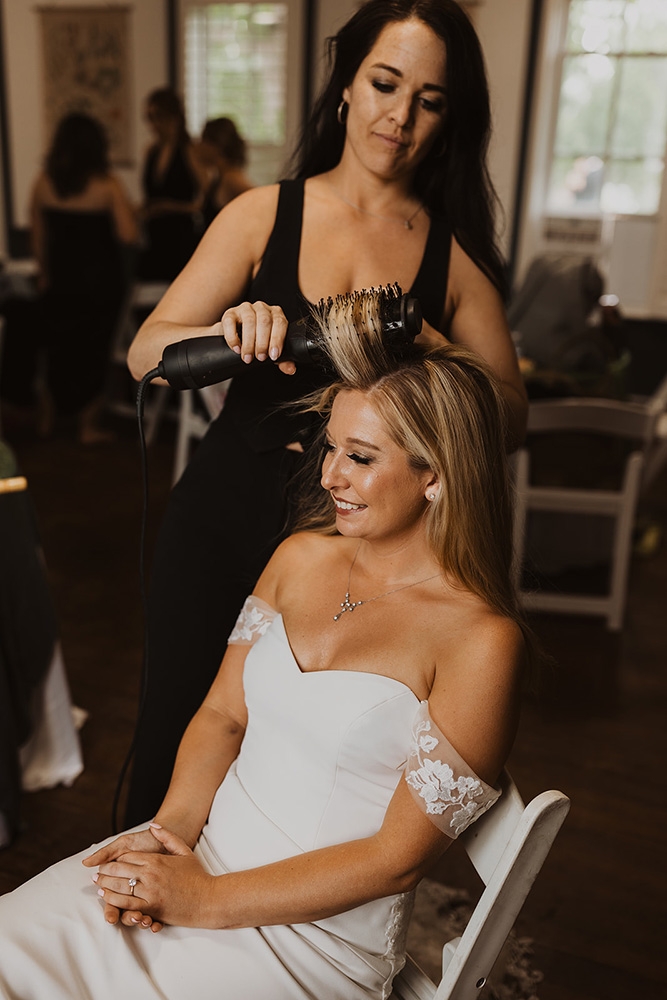 Standing hairdresser styling a sitting brides hair. The bride is smiling.  