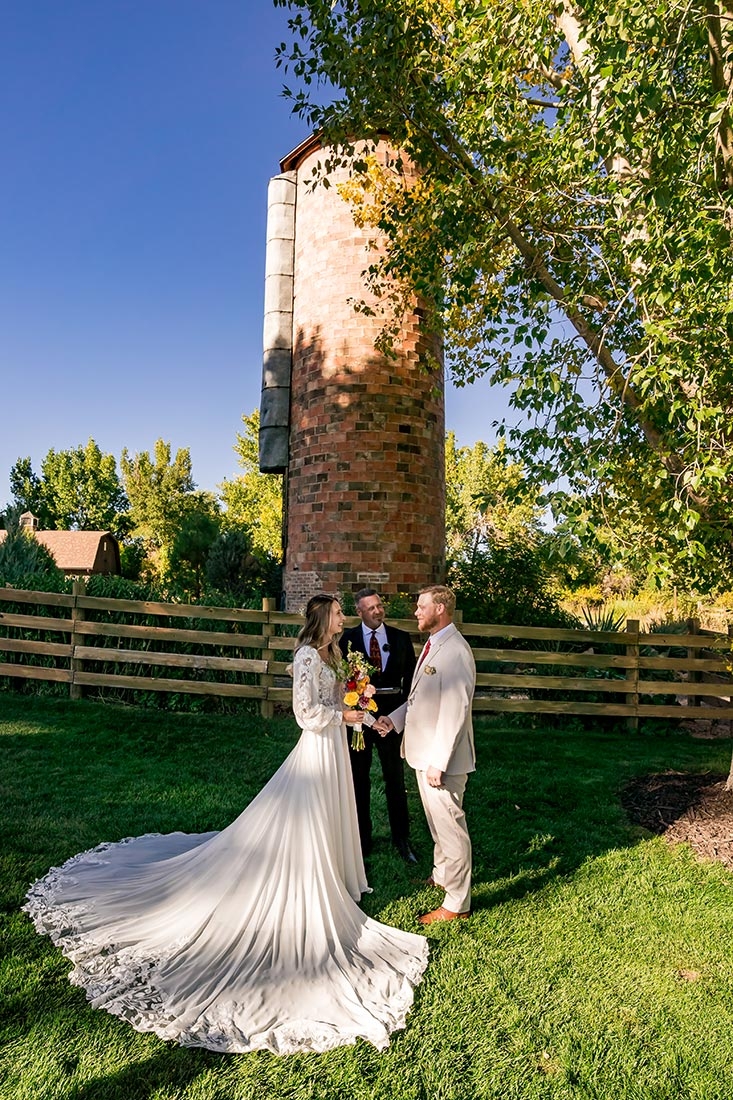 Bride and groom outside on a grassy area are with an officiant, standing in front of a silo.