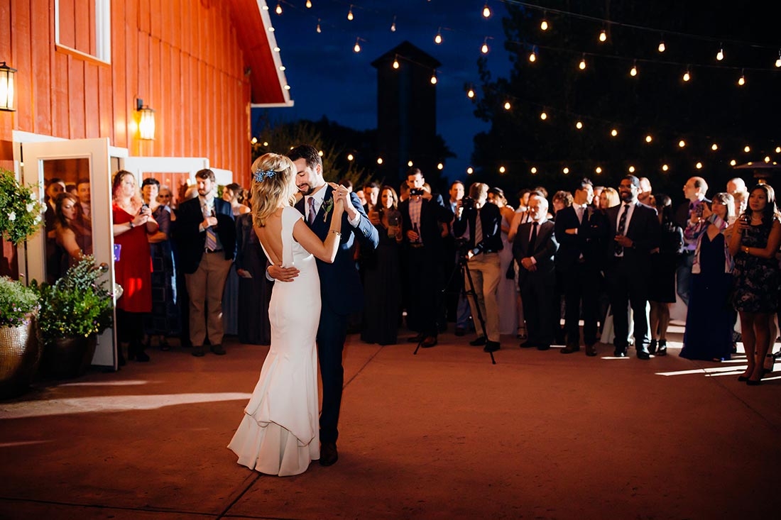 Bride and groom dancing outside of a barn at night under sparking lights with a crowd watching.