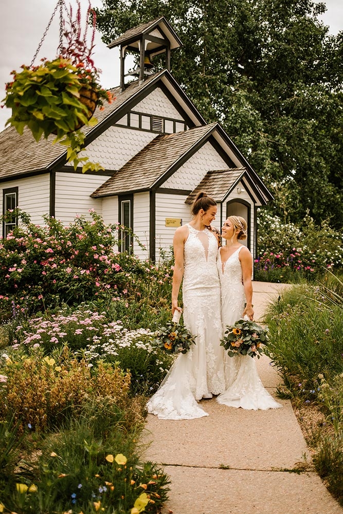 Two people in wedding dresses standing and smiling at each other while holding bouquets and standing outside in front of an old schoolhouse