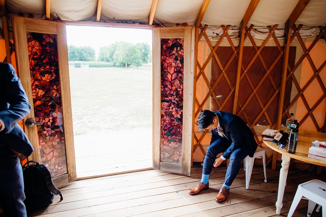 Man inside a yurt, sitting in a chair and leaning down, adjusting his pants leg. The door is open and you can see grassy fields in the background.