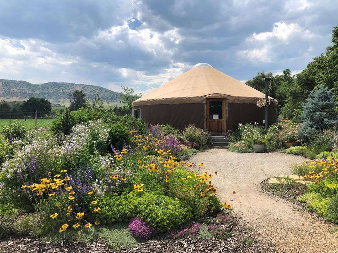 Yurt with colorful flowers in the foreground and mountains and blue sky with white clouds in the background. 