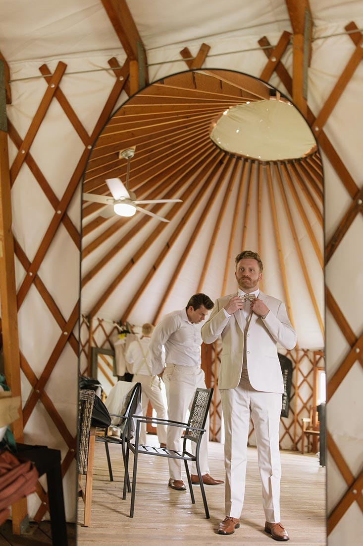 Groom getting ready for his wedding. He is inside of a yurt, looking into a mirror and adjusting his tie. 