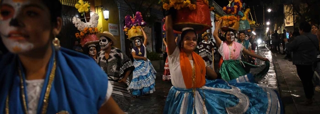 Women in brightly colored dresses walking through the streets of Oaxaca, Mexico at night with baskets of flowers balanced on their heads during the Día de los Muertos festival.