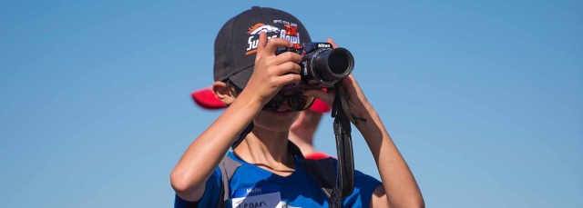 Child with blue shirt and ball cap looking through camera lens