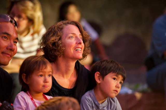 Family Programs Two adults and two children listening to a story. There are people behind them in a blurred image.
