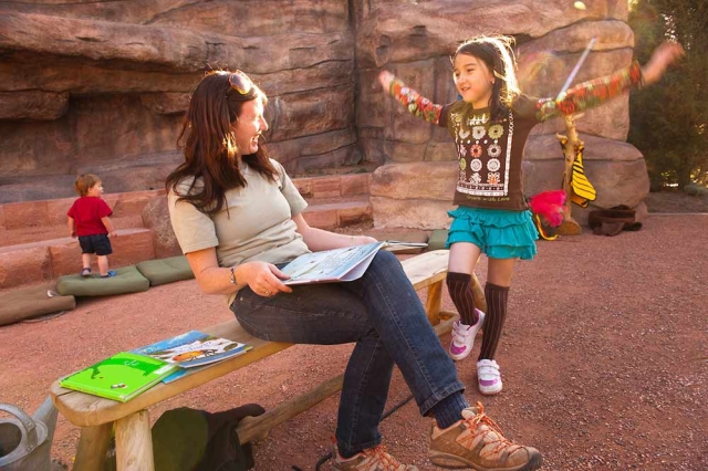 Family Programs Smiling seated adult outside looking at child standing with her arms outreached. The adult has an open book in her lap.