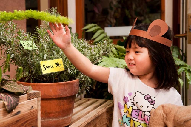 Family Programs Child wearing brown paper headband with cut out animal ears. They are touching a plant. The potted plant has a sign stuck into the dirt that says 'smell me'.
