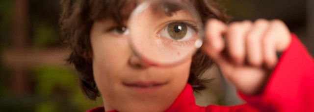 Child in red shirt looking through a magnifying glass. It is magnifying his left eye. 