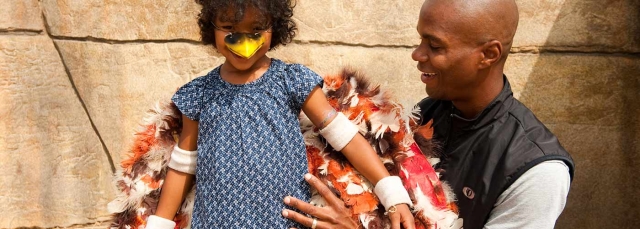 Adult holding a child wearing a feather costume and a toy beak.