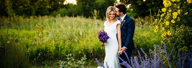 A bride and groom at their wedding, standing in an open field. She is holding a bouquet wit purple flowers. 