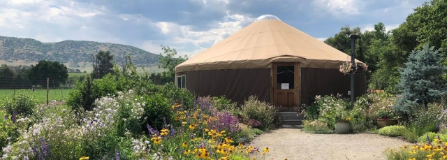 A yurt surrounded by colorful flowers, with mountains and a blue sky with white clouds in the background 