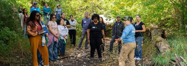 A guide talks to a group of people that are outside in a wooded area.