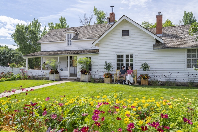 White farmhouse with pink and yellow flowers in the foreground. Two people are sitting on a bench in front of the farmhouse.