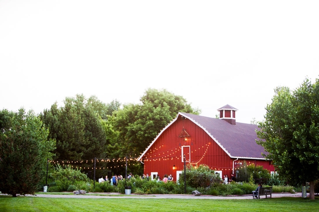 Red barn with green trees and green grass. There are twinkle lights hanging above a group of people.