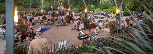 Group of people outside under a covered pergola are listening to a person playing a keyboard.